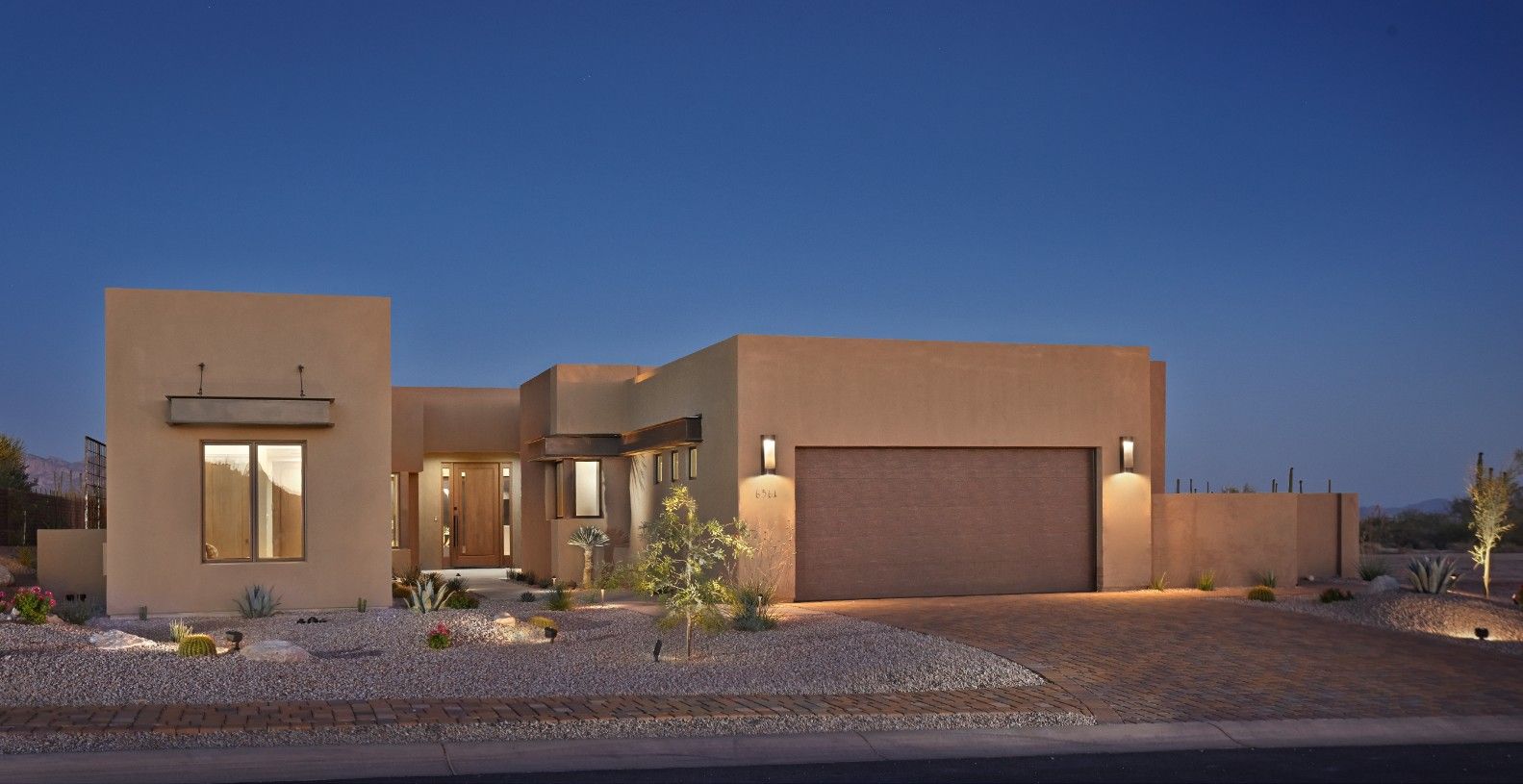 Contemporary adobe-style elevation at dusk with a lit garage and desert plantings