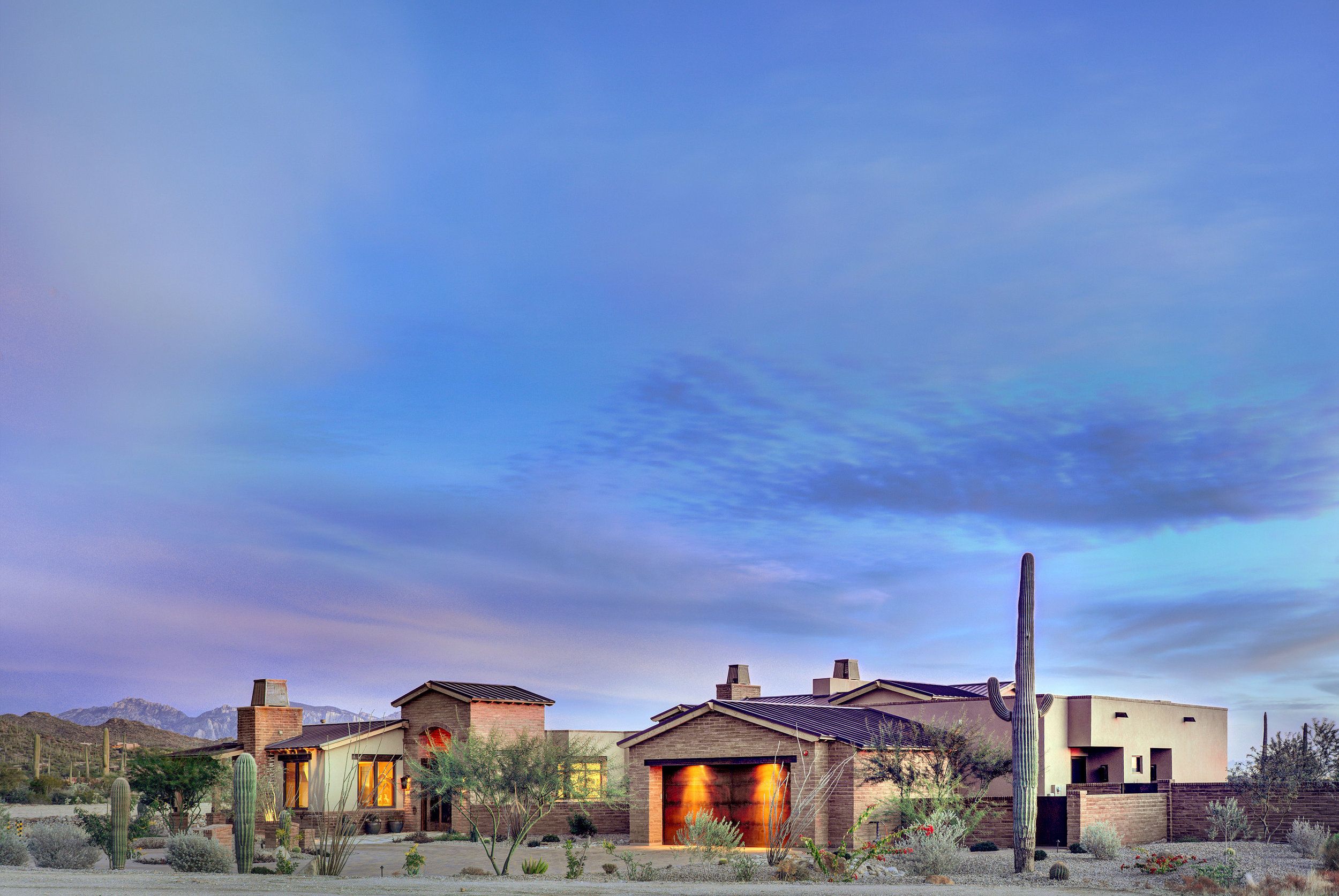 Single-storey territorial ranch home at dusk with lit windows and a saguaro against a wide desert sky