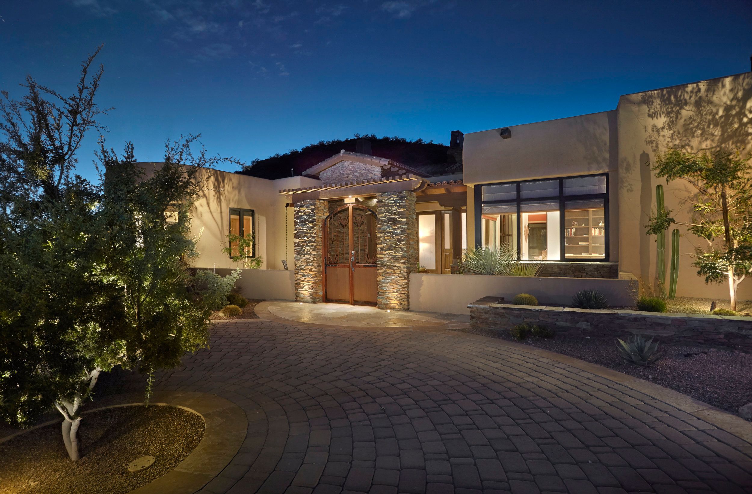 Lit entry and paver drive at twilight with mature desert trees
