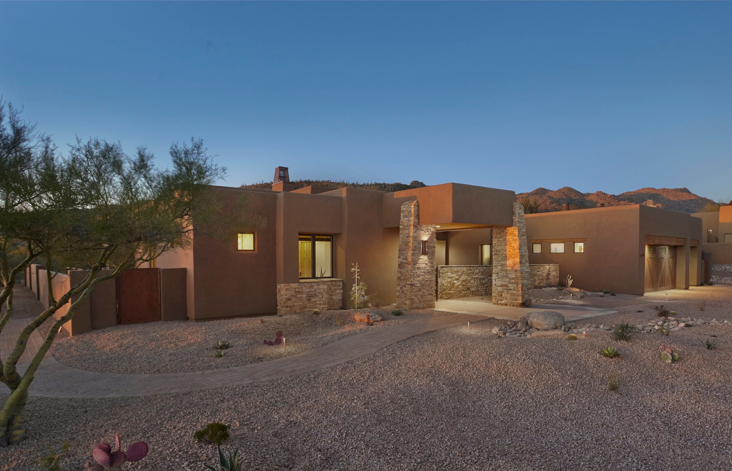 Low adobe elevation with stone piers and desert landscaping at dusk