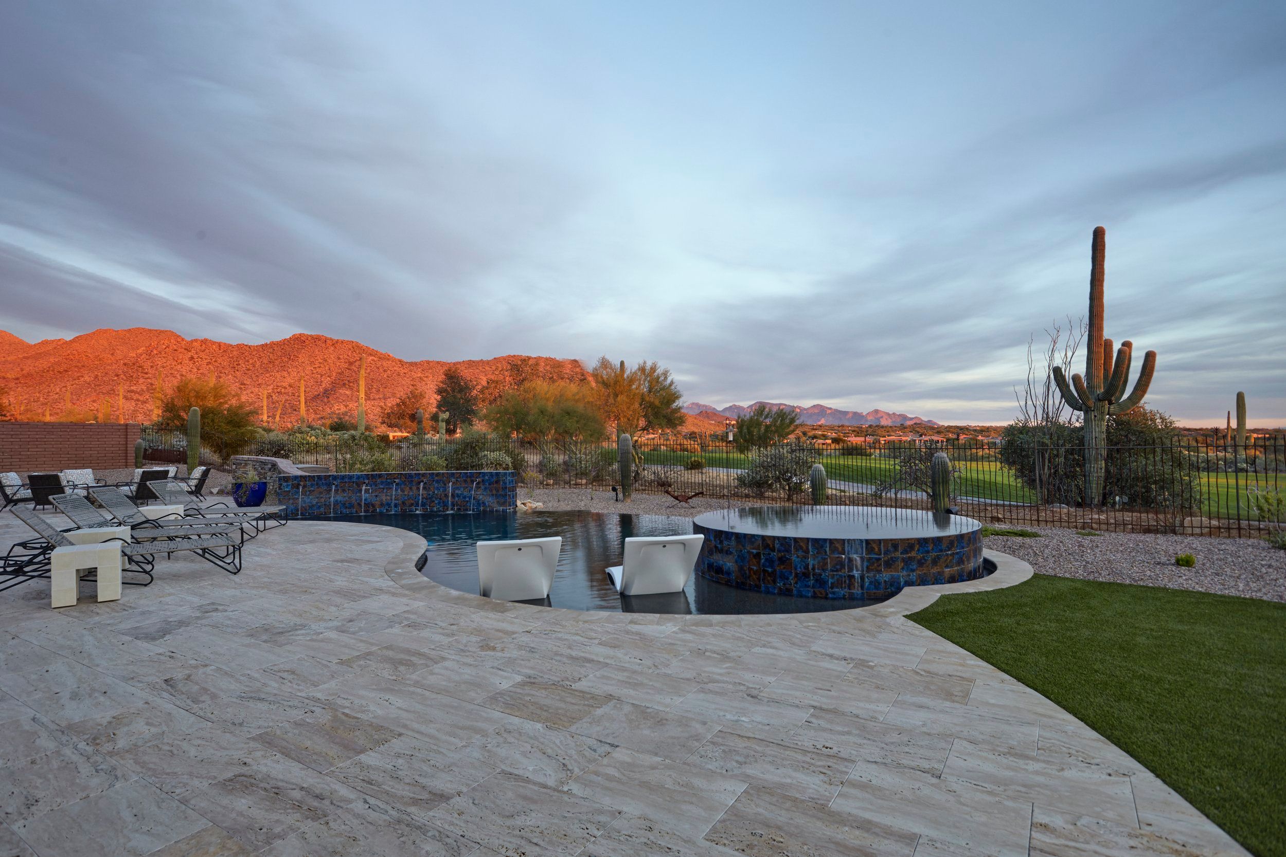 Pool and spa on a travertine deck with lounge chairs and saguaros against the mountains