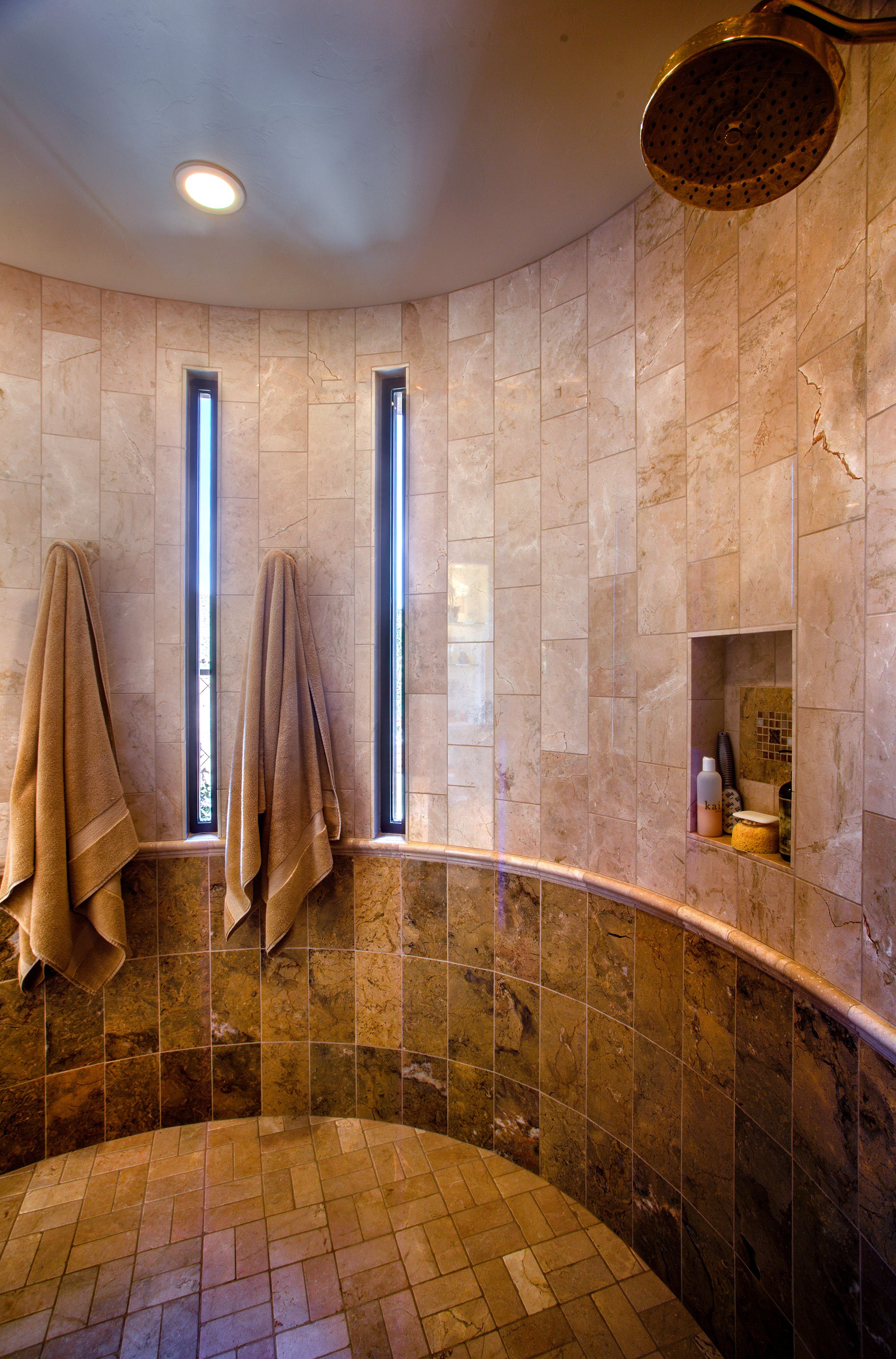 Curved walk-in travertine shower with a rain head and narrow slot windows