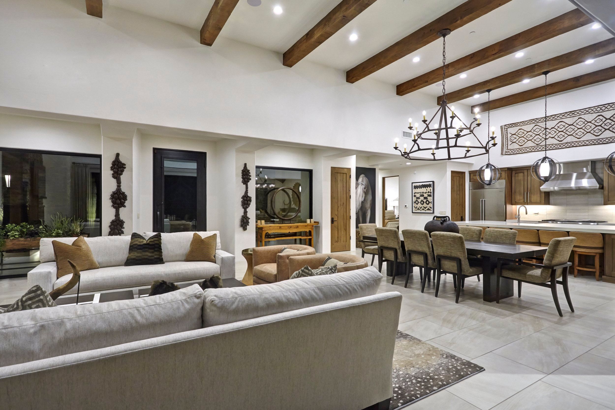 Open living and dining room with exposed beams, an iron chandelier and kitchen beyond