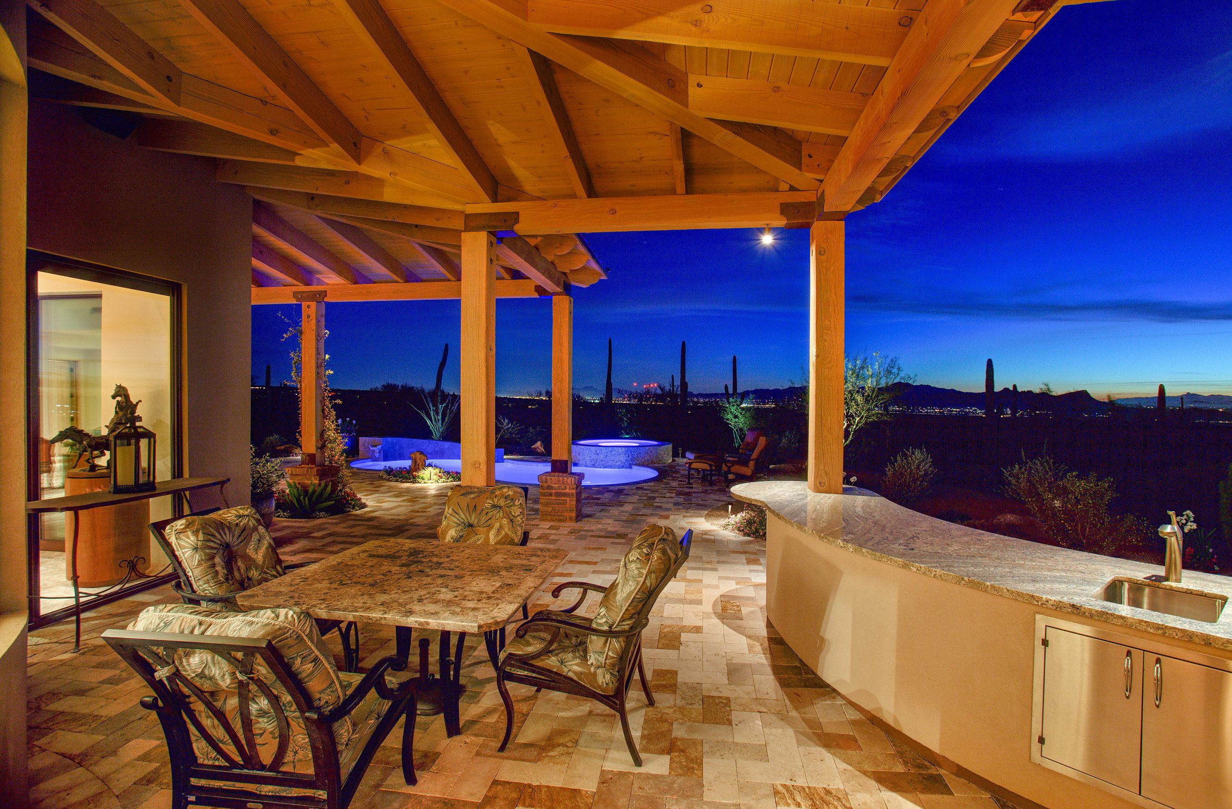 Covered patio at twilight with outdoor dining, a bar sink and lit pool beyond