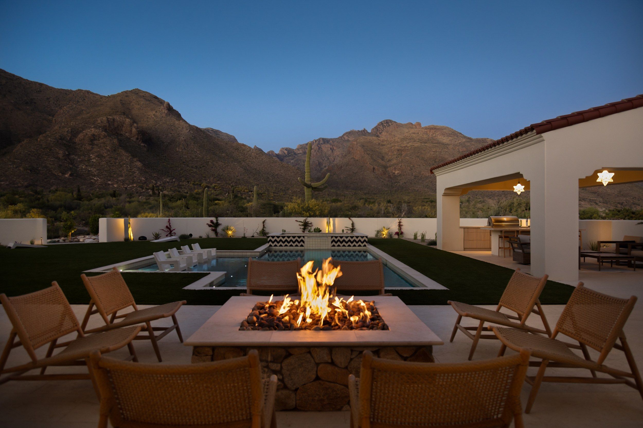 Fire table on the lawn at dusk with the pool, ramada and mountains behind